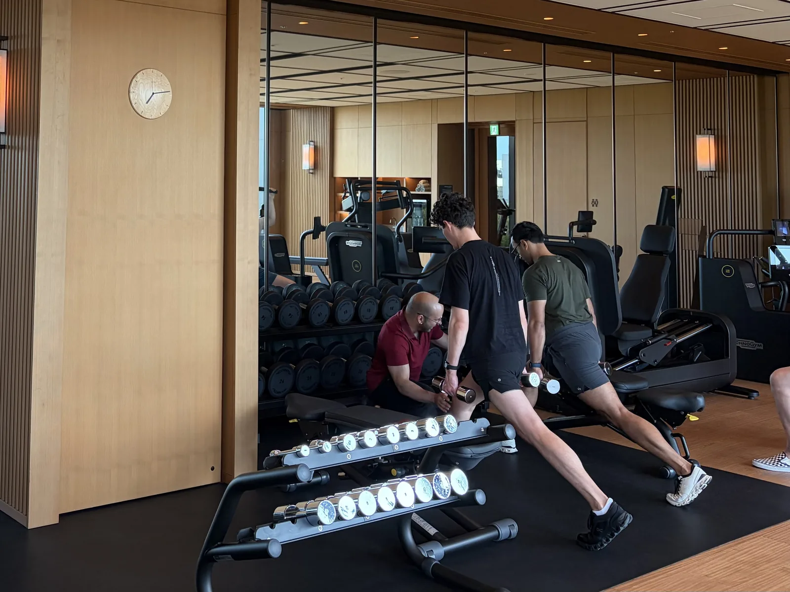 A trainer kneeling to check form while a guest performs a dumbbell lunge in a hotel gym