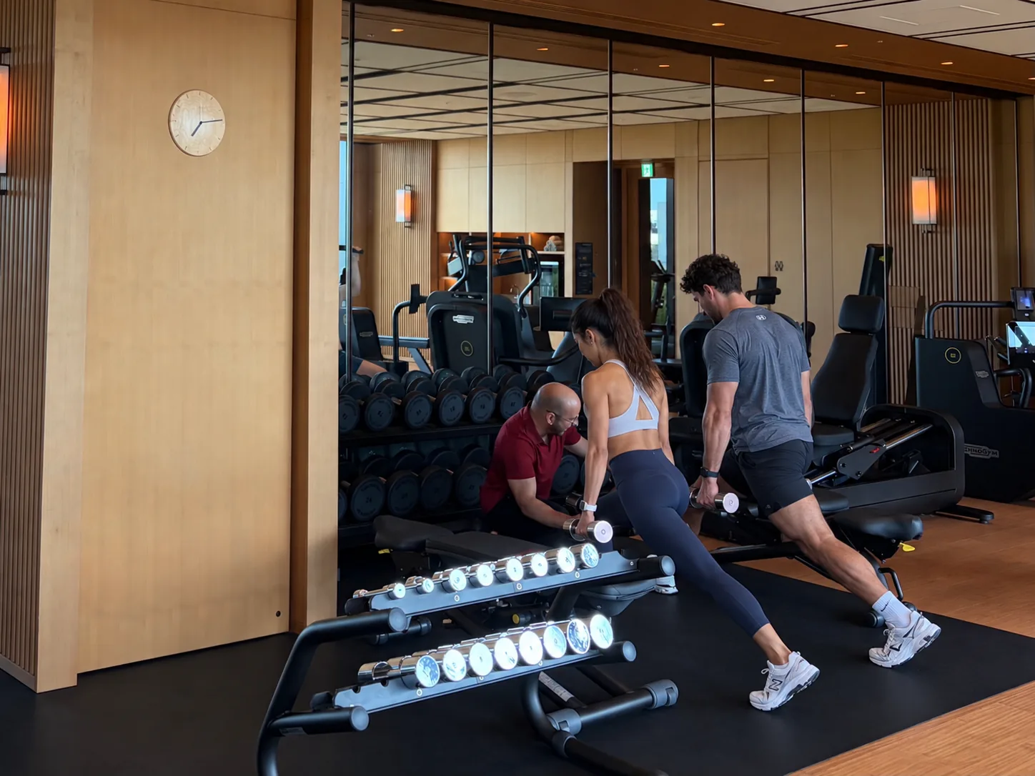 A FIT STAGE trainer kneeling to check form while two hotel guests perform dumbbell lunges in a hotel gym in Osaka