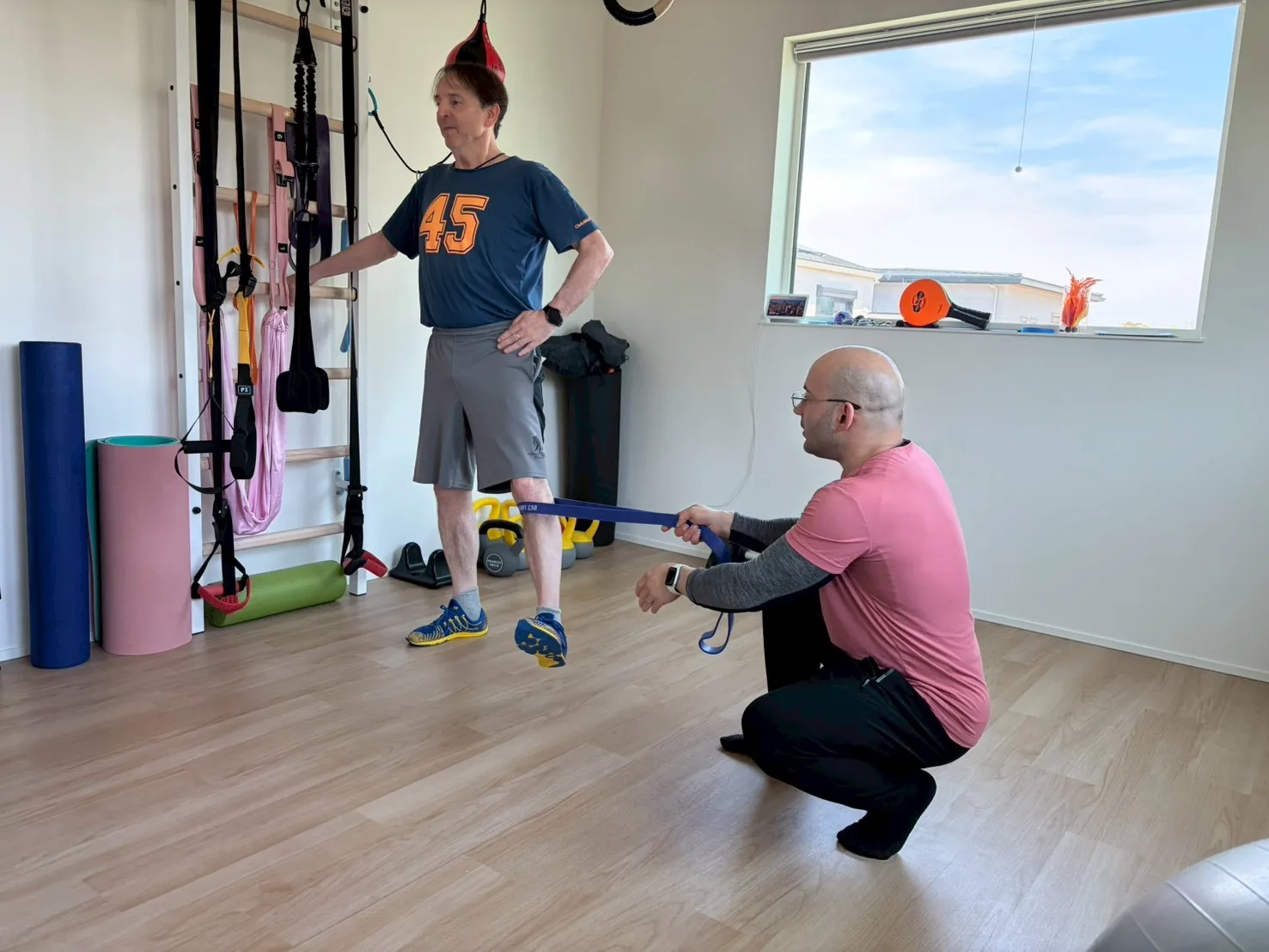 English-speaking personal trainer coaching an older client through a single-leg balance exercise with a resistance band in a bright private training studio