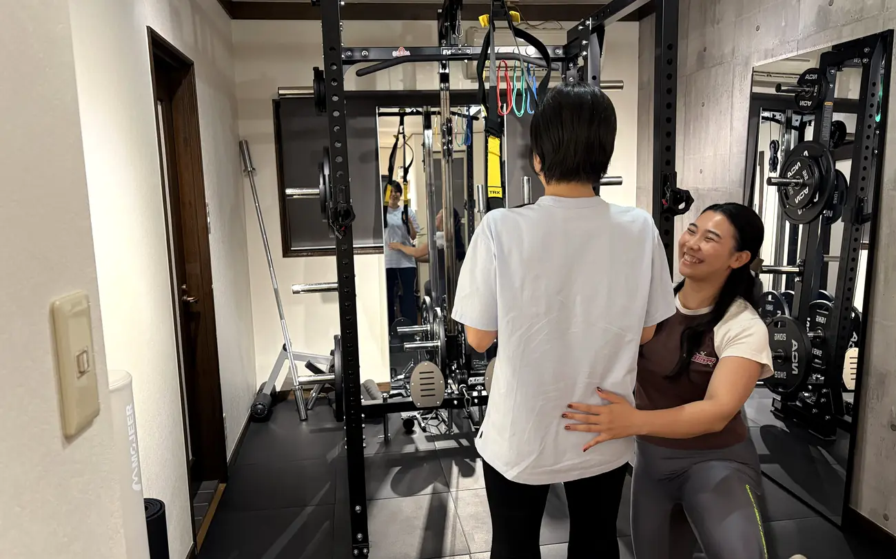 ラックの前で、トレーナーが腰に手を添えて立ち姿勢を確認する様子 — FIT STAGE江坂店