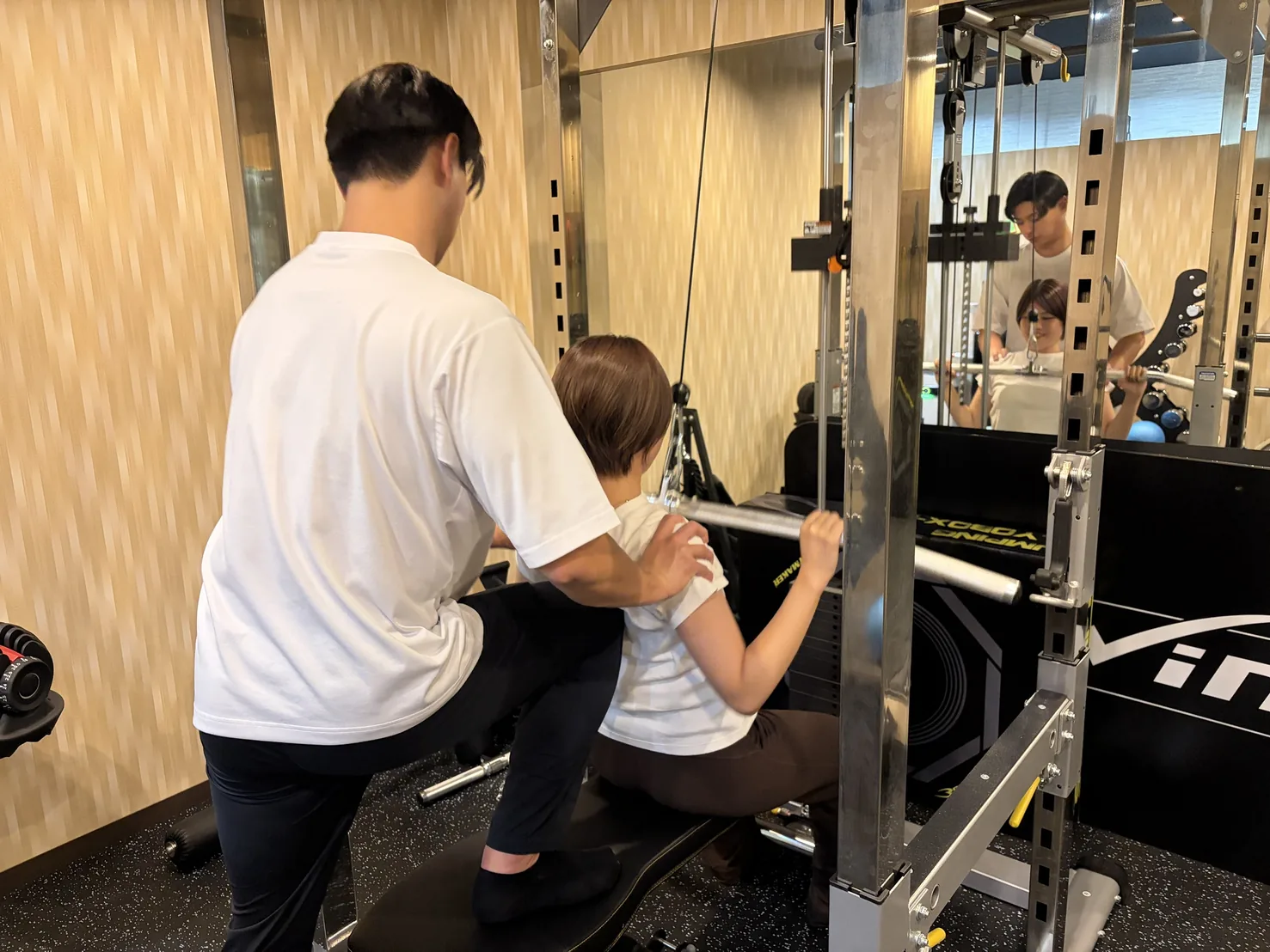 FIT STAGE 庄内店で、ラックのバーを担ぐ動作をトレーナーが肩に手を添えて支える様子