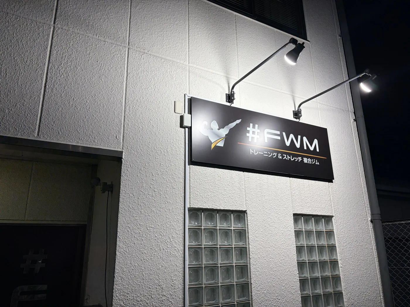 夜にライトアップされたFIT STAGE 庄内店の建物壁面看板「#FWM トレーニング＆ストレッチ 複合ジム」